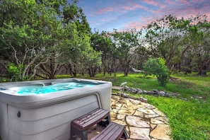 A hot tub with a stone patio and a wooden bench, surrounded by trees and a grassy area.