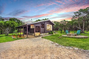 A rustic cabin with a covered porch, a stone fireplace, and a view of the surrounding greenery. - Canyon Vista I Romantic, hot tub, log cabin (Wimberley)