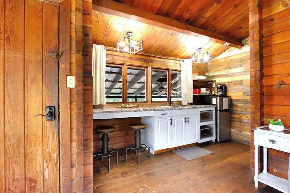 A kitchen with wooden walls, a countertop, a sink, a refrigerator, and a bar stool. - Canyon Vista I Romantic, hot tub, log cabin (Wimberley)