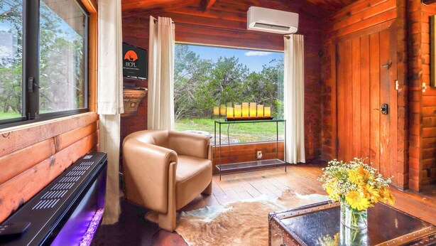 A cozy cabin interior with a leather chair, a piano, a glass table with a vase of flowers, and a view of the outdoors through large windows. - Canyon Vista I Romantic, hot tub, log cabin (Wimberley)