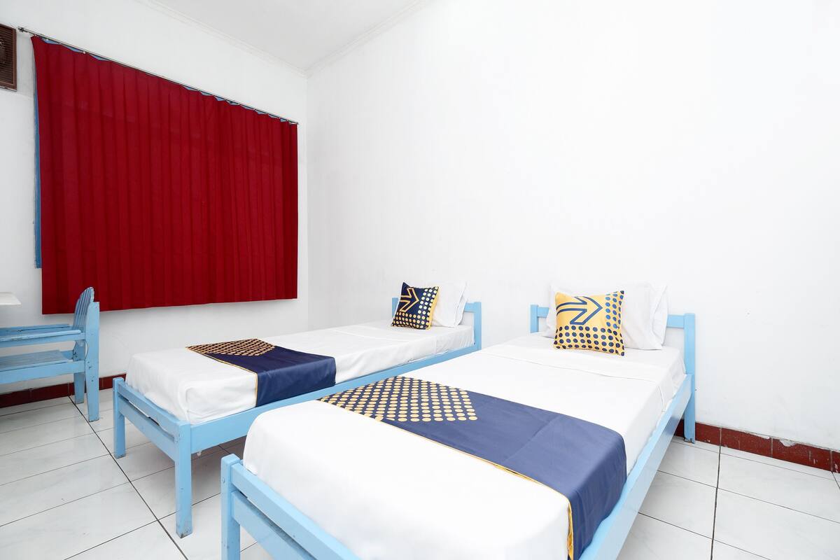 Quarto com duas camas de solteiro, cada uma com estrutura azul e roupa de cama branca. Há uma cortina vermelha na janela, e uma pequena mesa com uma cadeira à esquerda.