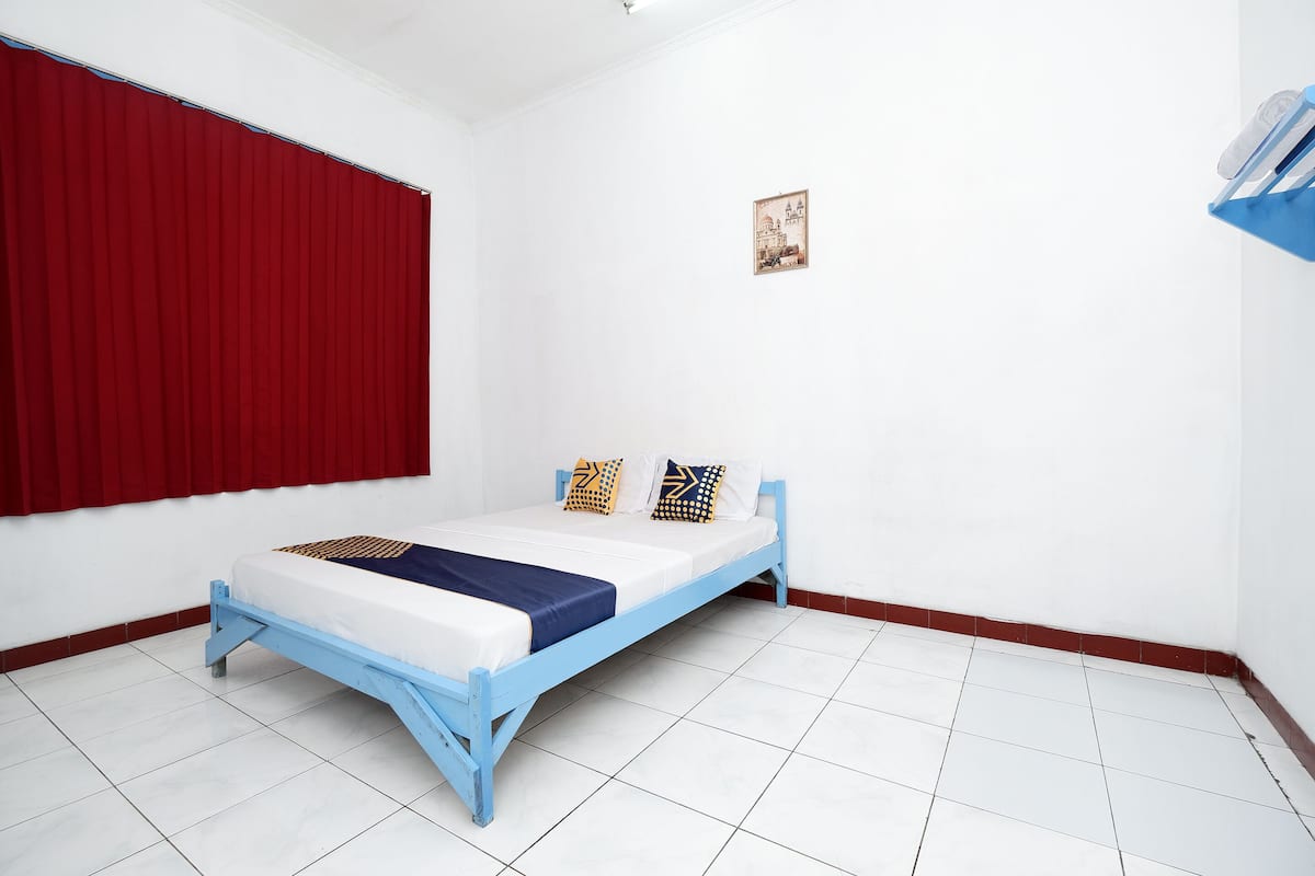 Um quarto com uma cama, cortinas vermelhas, um pequeno quadro na parede e um chão coberto de azulejos brancos.