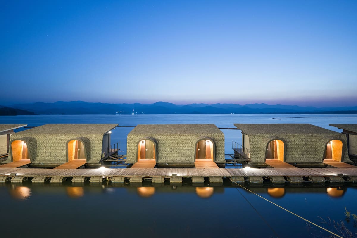 A row of overwater bungalows with wooden walkways and stone walls, connected by a bridge, overlooking a calm body of water and distant mountains.