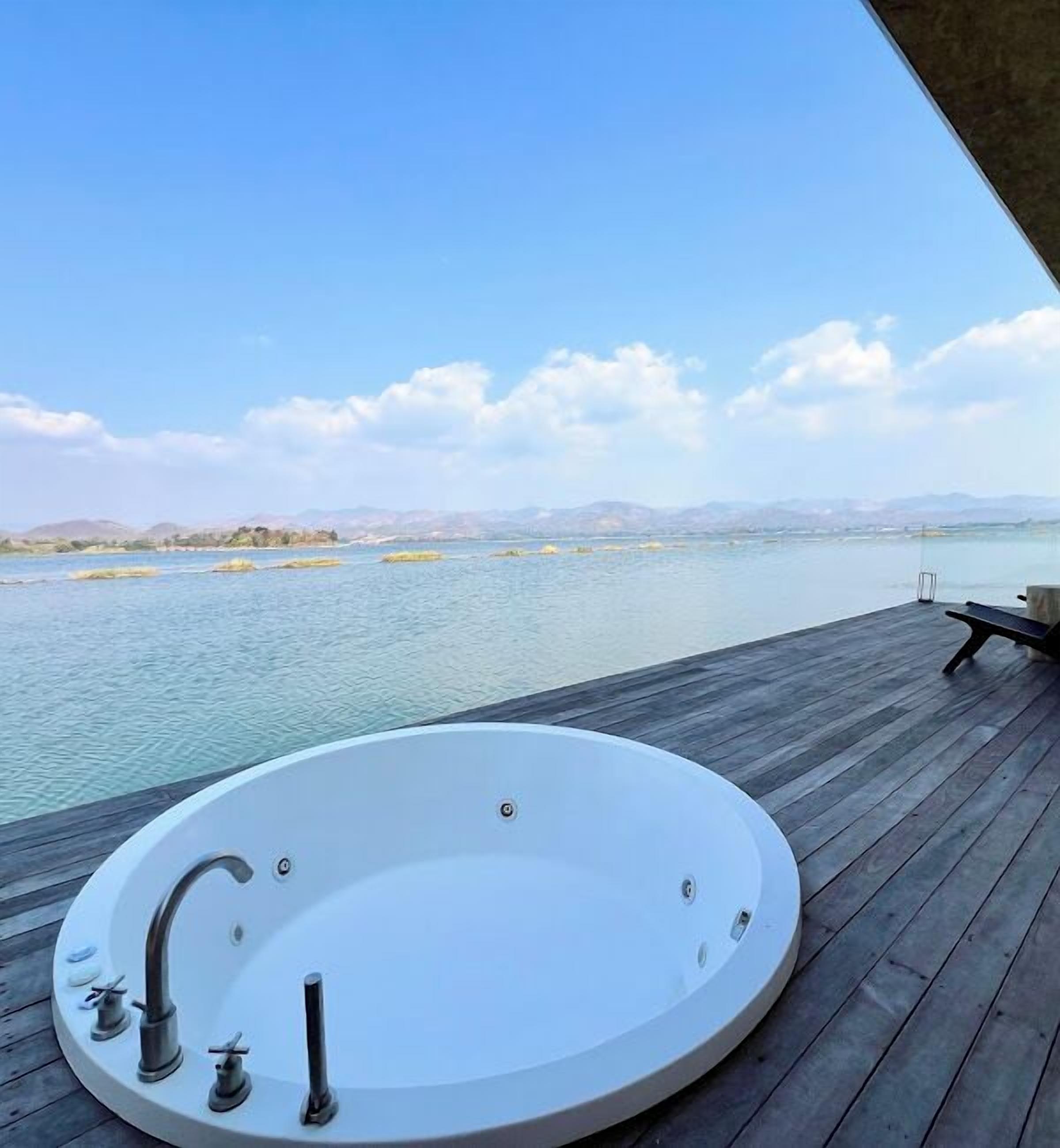 A circular hot tub on a wooden deck with a view of a calm body of water and distant mountains.