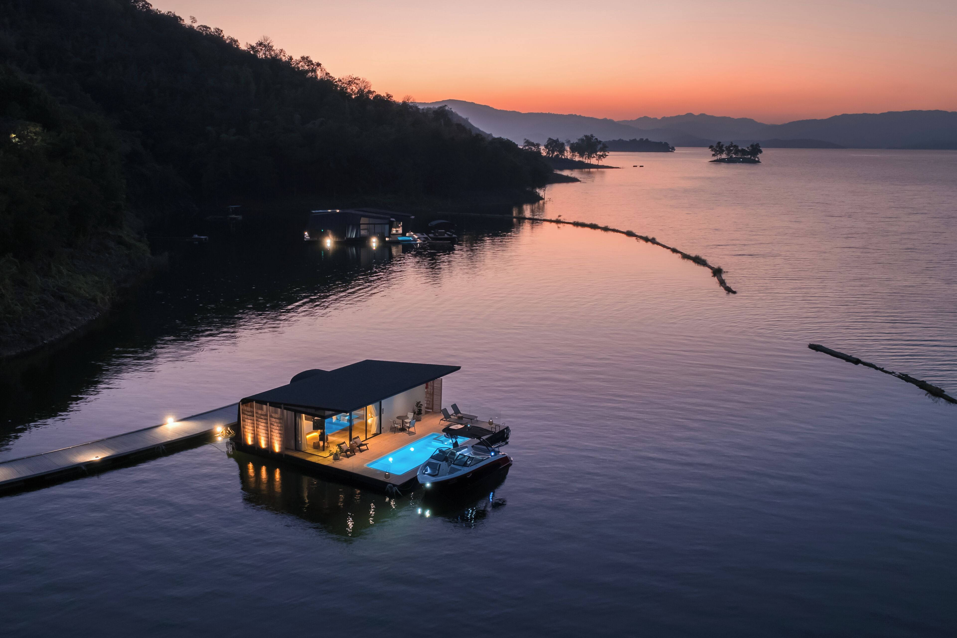 A floating house with a pool, docked boats, and a serene lake at sunset.