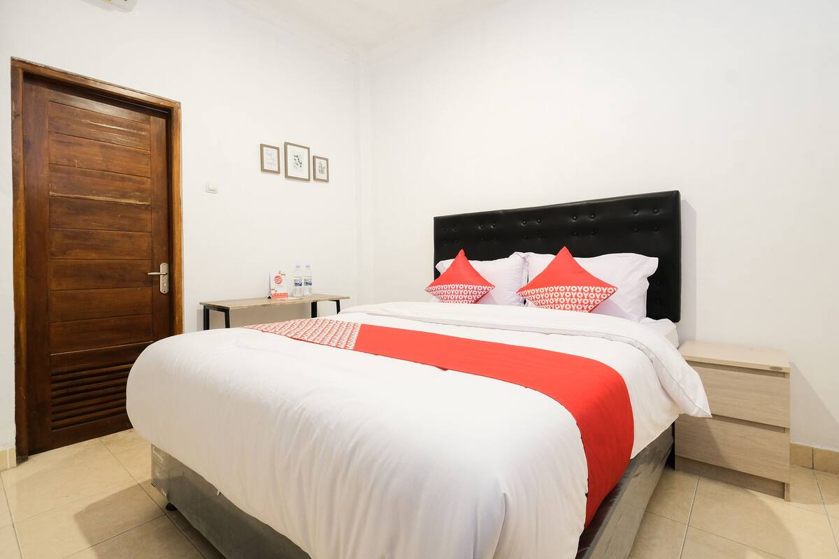 A neatly made bed with a white and red bedspread, a wooden door, and a small bedside table with a bottle and a glass.