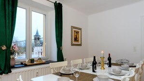 A dining room with a table set for four, white chairs, a window with green curtains, a radiator, and a framed picture on the wall. - Zythogite Appartement 9 personnes (Tintigny)
