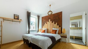 A bedroom with a wooden floor, a bed with white and brown bedding, a wooden headboard, a bedside table with a lamp, and a window with curtains. - Zythogite Appartement 9 personnes (Tintigny)