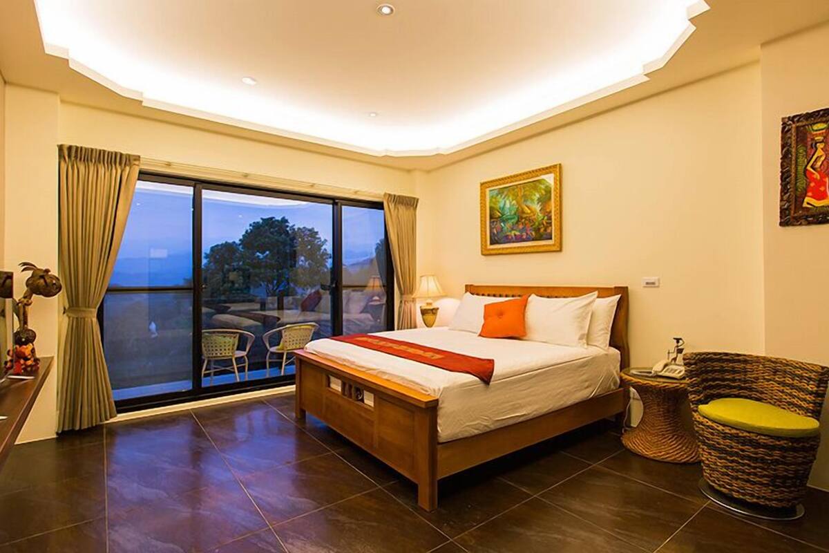 Un cuarto de hotel con una cama grande, vistas al exterior a través de una puerta corrediza, un cuadro en la pared y una silla de mimbre con cojín.