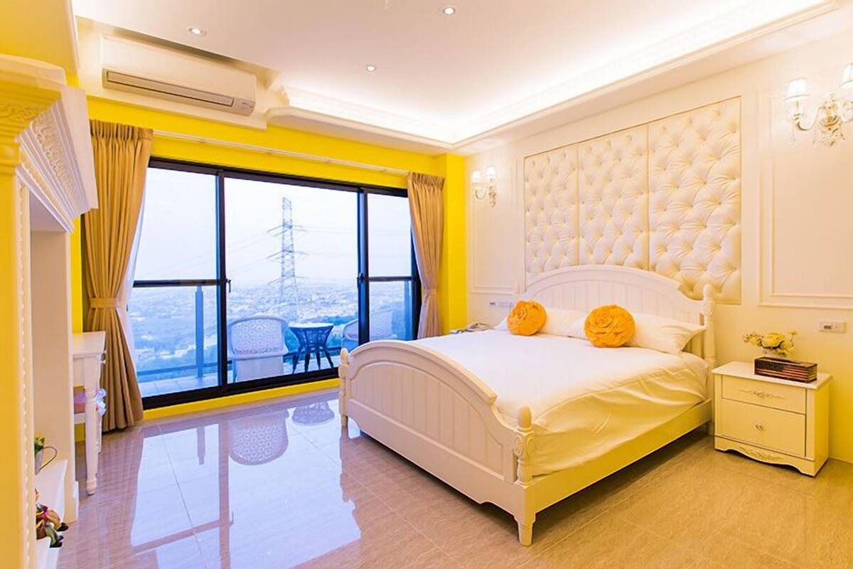 Un dormitorio con una cama grande, una mesita de noche, una silla y vistas de la ciudad a través de amplios ventanales.