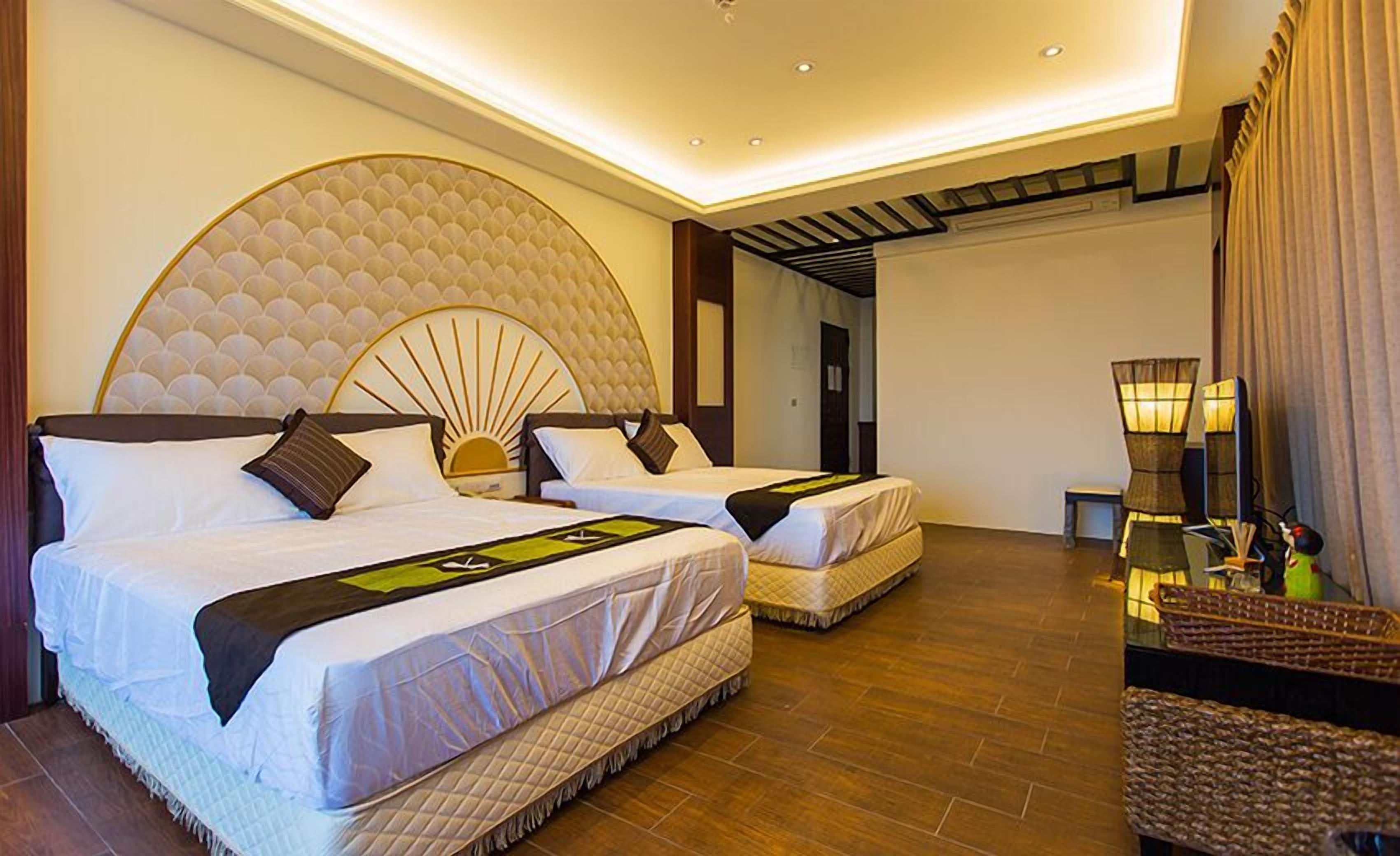 Une chambre d’hôtel avec deux lits, une grande tête de lit décorative, un plancher en bois et un petit coin salon avec une lampe.