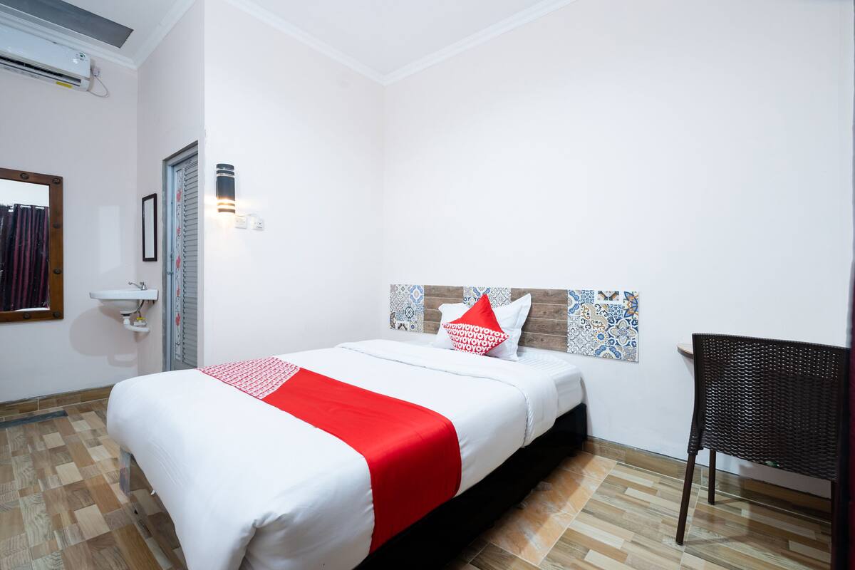 A hotel room with a bed, a chair, a wall-mounted shelf, and a patterned headboard.