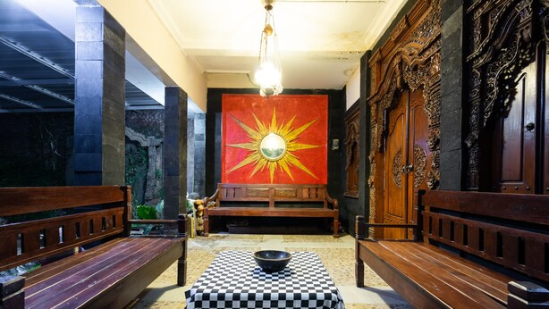 A room with a checkered table, wooden benches, a large mirror with a sun design, and ornate wooden doors. - Hotel O Art Guest House Syariah (Yogyakarta)