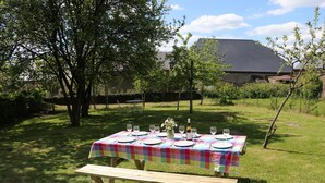 Una mesa de picnic montada en un césped, con un mantel colorido, rodeada de árboles y una casa al fondo.