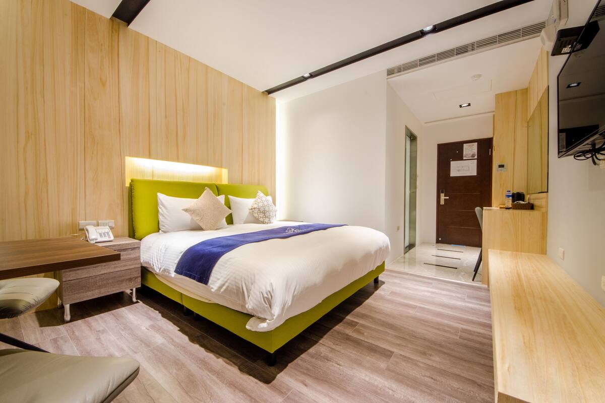 A modern hotel room with a green bed, white bedding, a bedside table with a phone, and a desk with a chair.