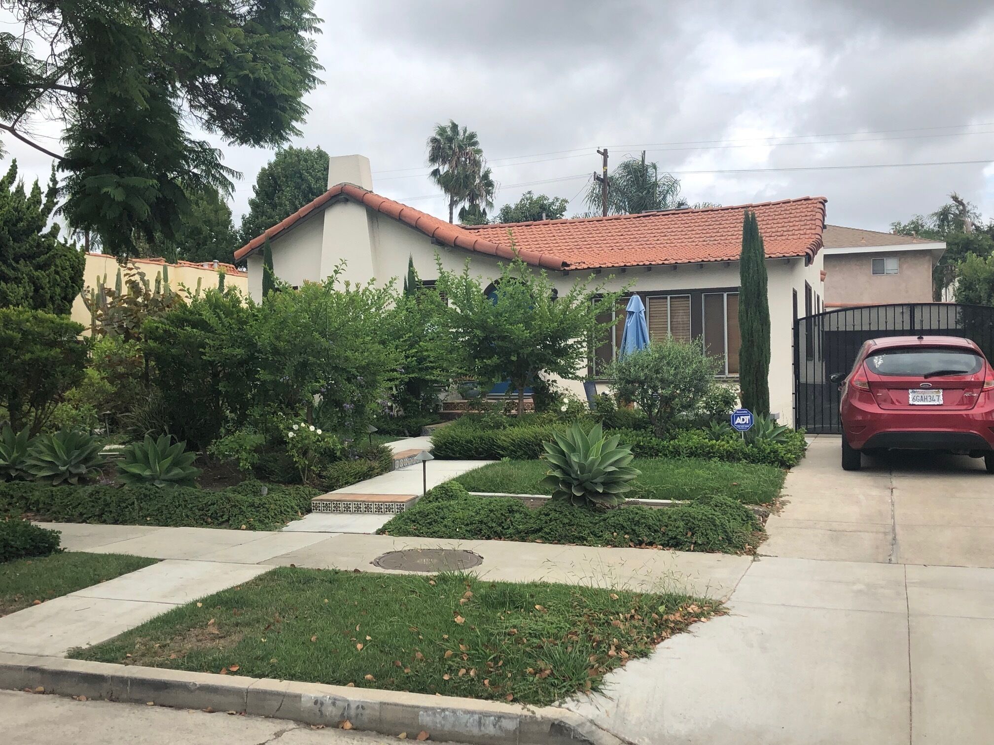 A residential area with a house featuring a red-tiled roof, a gated driveway, and a red car parked in the driveway.