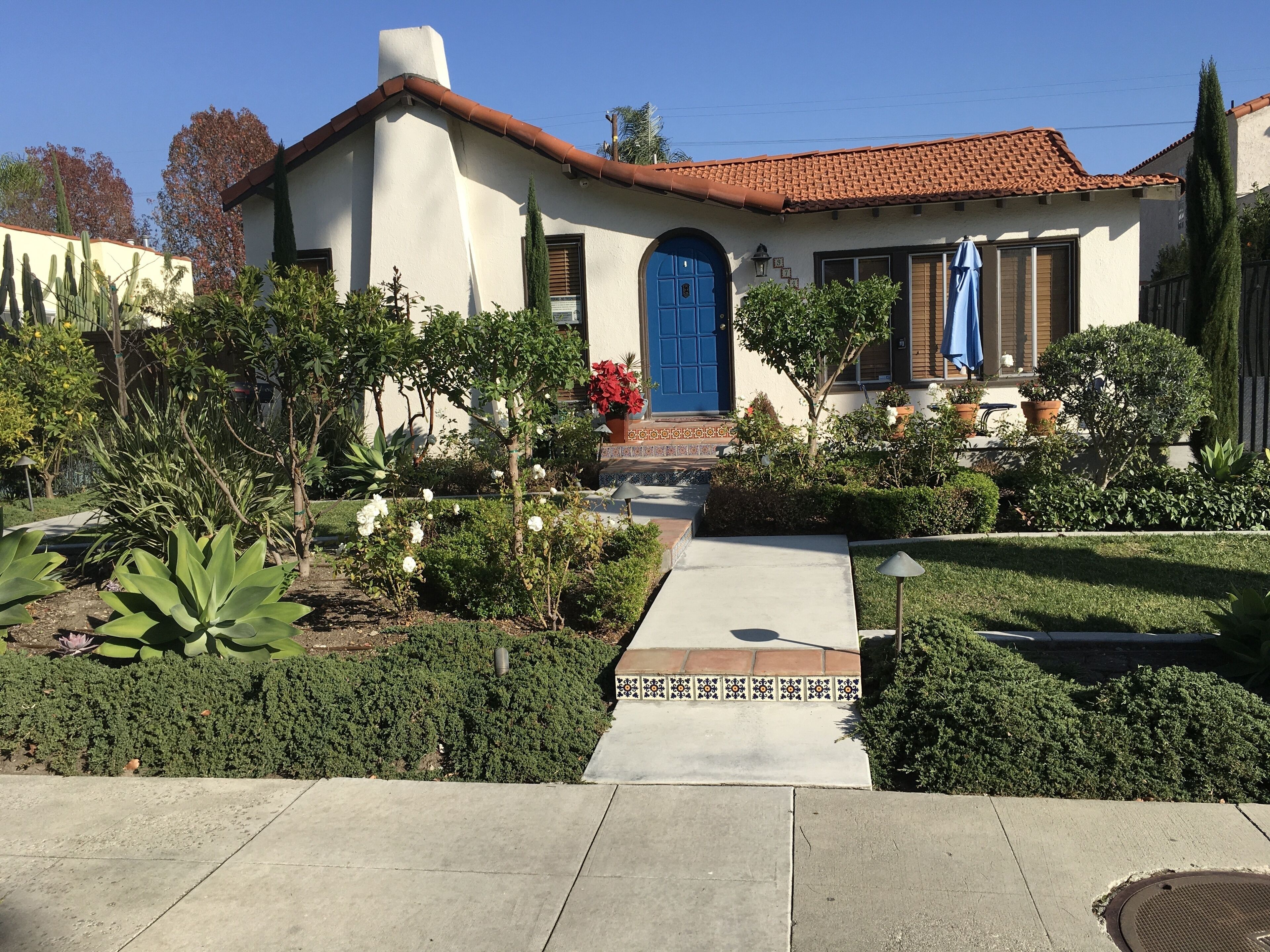 Ein Haus mit rotem Ziegeldach, blauer Tür und einem gepflegten Garten mit verschiedenen Pflanzen und einem Weg zum Eingang.