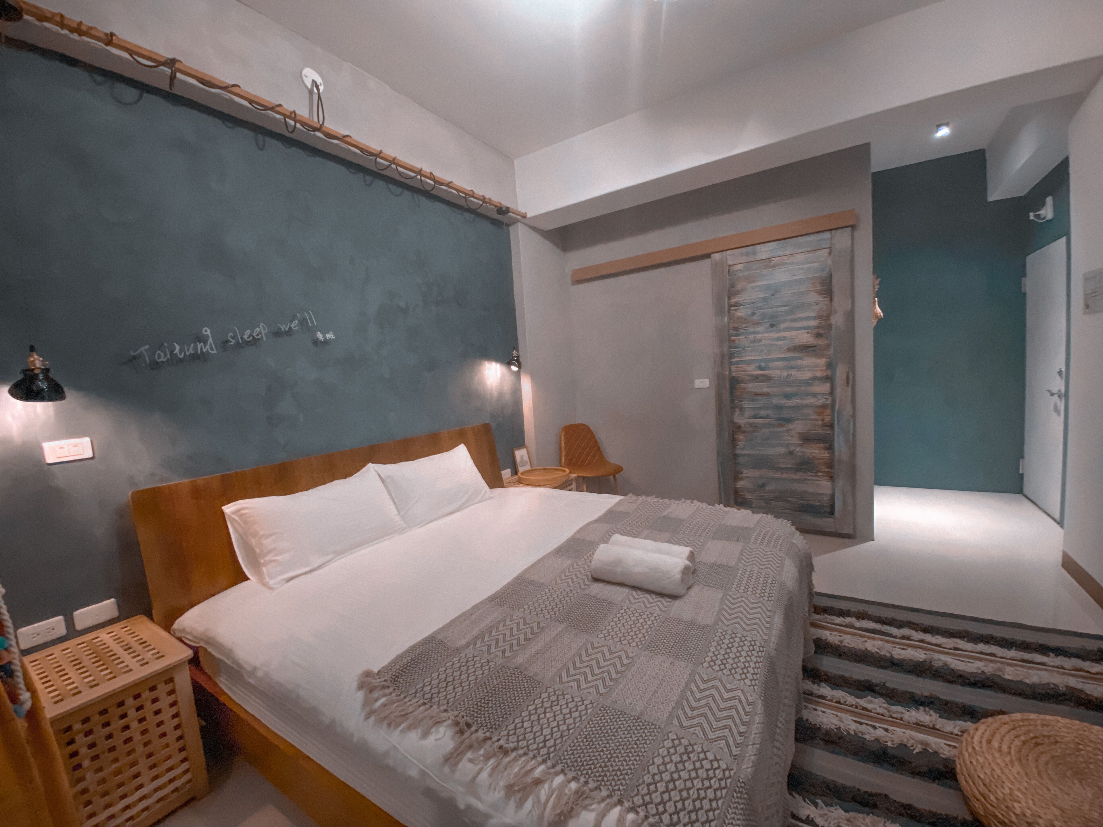 Un dormitorio moderno con una cama grande, cabecera de madera, estante en la pared, una mesita y una silla.