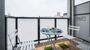 Un balcon avec une table dressée pour deux personnes, des chaises blanches et un petit coin jardin agrémenté de galets blancs et d’herbe verte.