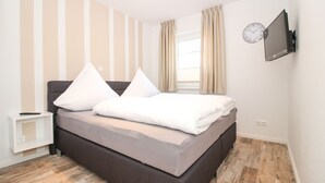Un lit bien fait, avec des oreillers blancs et un matelas gris, dans une pièce dont le papier peint est à rayures, avec une fenêtre ornée de rideaux et un téléviseur fixé au mur.