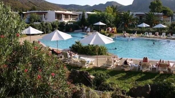 Ein Poolbereich einer Ferienanlage mit mehreren Liegestühlen und Sonnenschirmen, umgeben von üppiger Vegetation und einer Bergkulisse.
