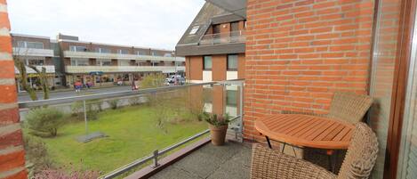 A balcony with wicker furniture, a potted plant, and a view of a green lawn and buildings in the background.