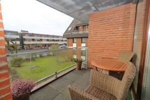 Ein Balkon mit Korbmöbeln, einem Blumentopf und Blick auf eine grüne Rasenfläche sowie Gebäude im Hintergrund.