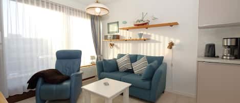 Una sala de estar con un sofá azul, una mesa de centro blanca, un sillón azul y una zona de cocina con una cafetera y un hervidor.