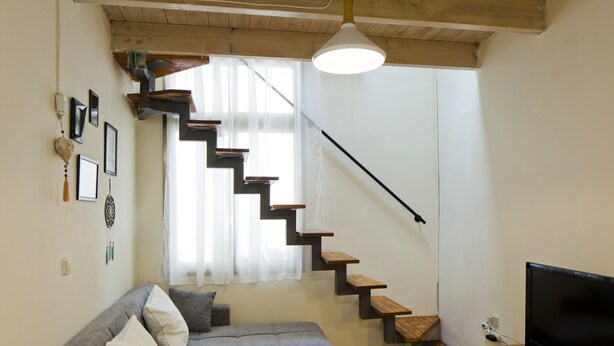 A modern living room with a grey sectional sofa, a wooden coffee table, a flat-screen TV, a staircase with wooden steps, and a large window with sheer curtains. - Stylish flat at Rothschild boulevard (Tel Aviv)