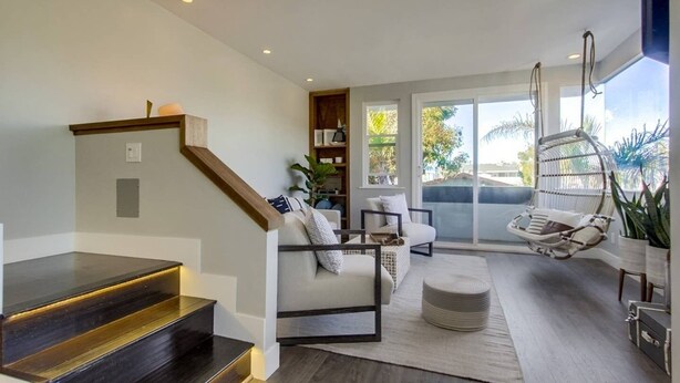 A modern living room with a staircase, a sofa set, a hanging chair, and a large window with a view of the outdoors.