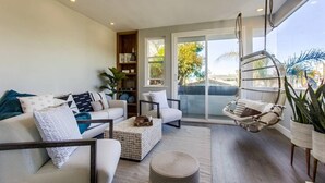 A modern living room with a sofa, coffee table, and hanging chair. There is a sliding glass door leading to a balcony with a view of trees and buildings.