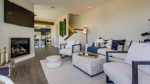 A modern living room with a fireplace, a flat-screen TV mounted on the wall, a sofa set, a coffee table, and a staircase leading to the kitchen.