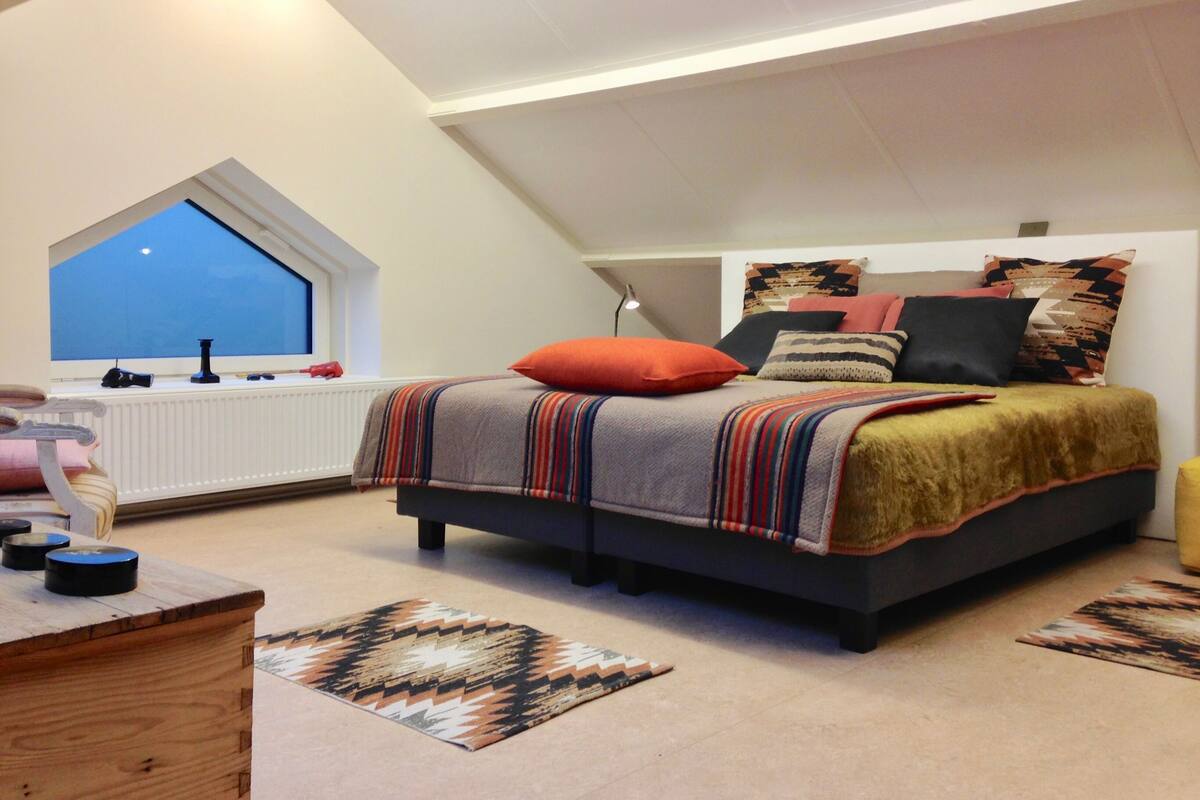 A bedroom with a sloped ceiling, a bed with multiple pillows, a window, and a radiator.