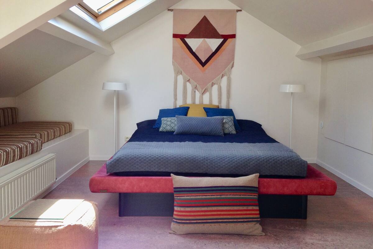 A bedroom with a sloped ceiling, a bed with a blue cover, a red bench, a striped pillow, and a geometric wall hanging.