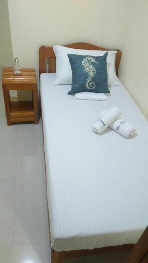 A single bed with white bedding, a teal pillow with a seahorse design, and two rolled towels with text on them.