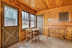 Relax in this serene space with a large window offering views of the surrounding trees, a wooden bench, and a dining table with chairs.