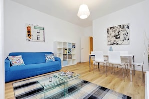 Uma sala de estar moderna com um sofá azul, mesa de centro de vidro, área de jantar branca e uma parede com o letreiro 'Broadway'.