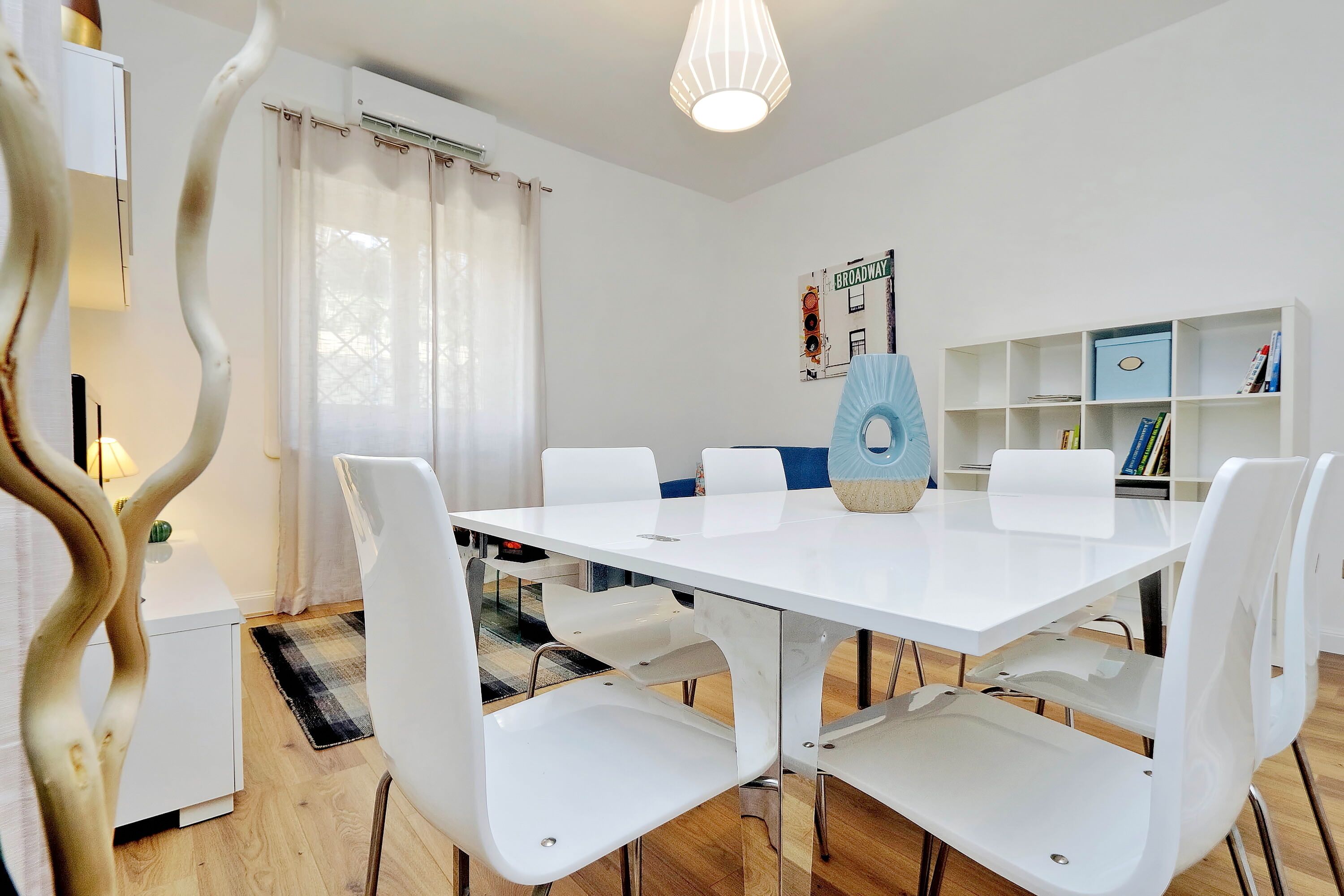 A modern dining area with a white table and chairs, a blue decorative item on the table, and a bookshelf with books and a blue box.