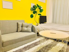 A room with a sofa, a round table, a bed, a TV, and a plant.
