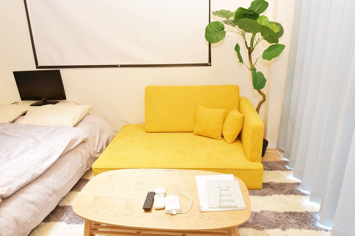 A hotel room with a yellow sofa, a bed with white bedding, a small wooden table with a remote control, a power strip, and a plant.