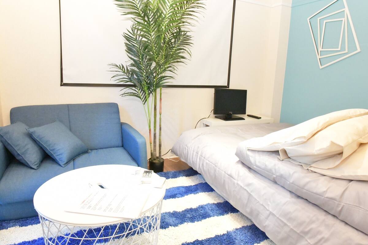A room with a blue sofa, a white round table, a bed with white bedding, a blue and white striped rug, and a potted plant.