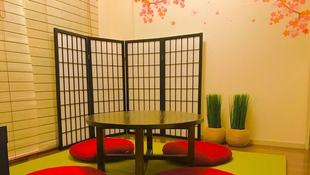 Een kamer met een tatami vloer, een lage tafel met zwart frame en rode kussens. Twee potplanten en een decoratieve muur met kersenbloesems.