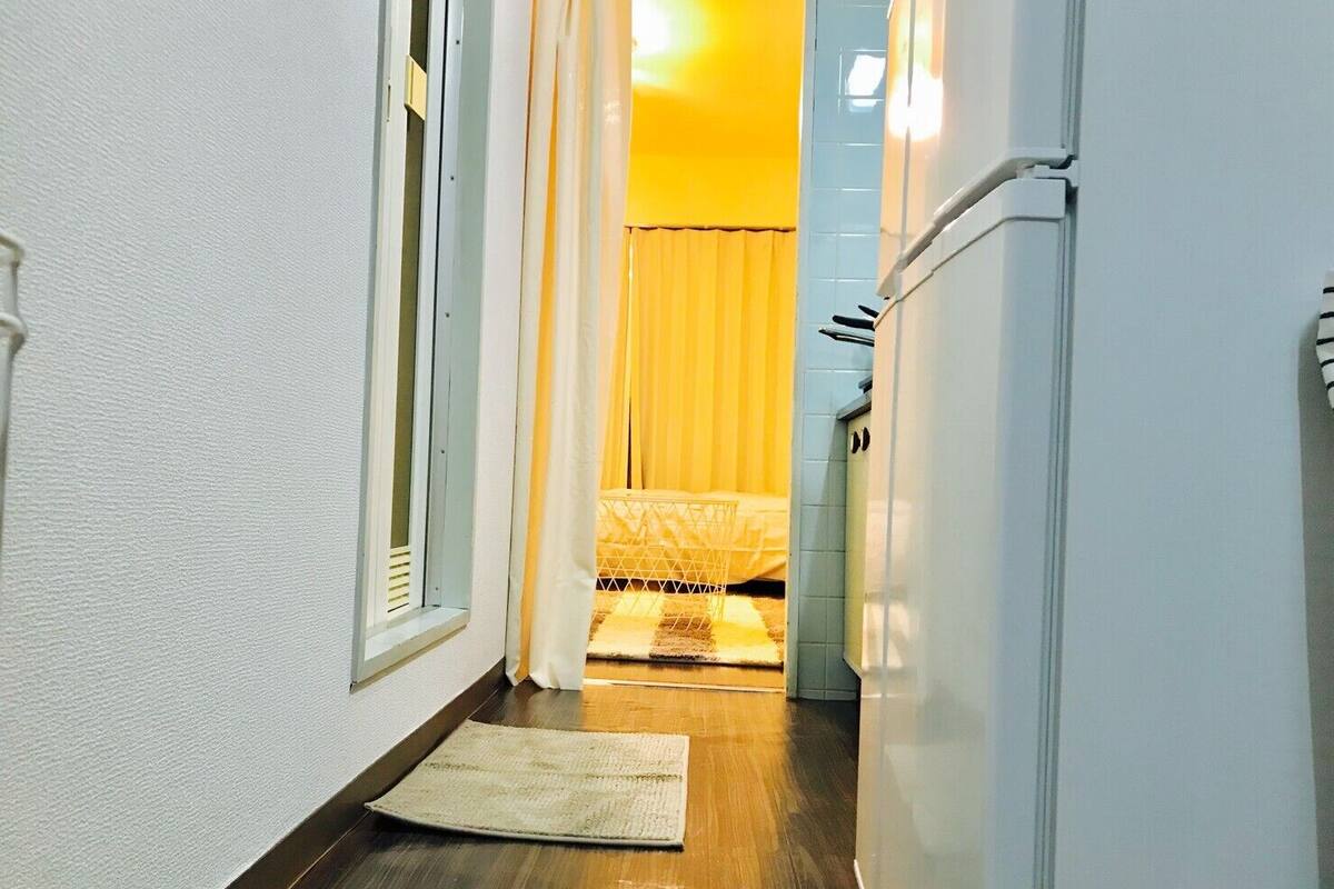 A narrow hallway with a wooden floor, a white door on the right, and a bed with a yellow curtain in the background.