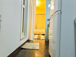 A narrow hallway with a wooden floor, a white door on the right, and a bed with a yellow curtain in the background.