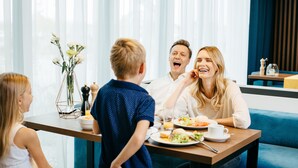 En familj sitter vid ett bord och äter. På bordet står en vas med blommor, en skål med mat och ett glas vatten.