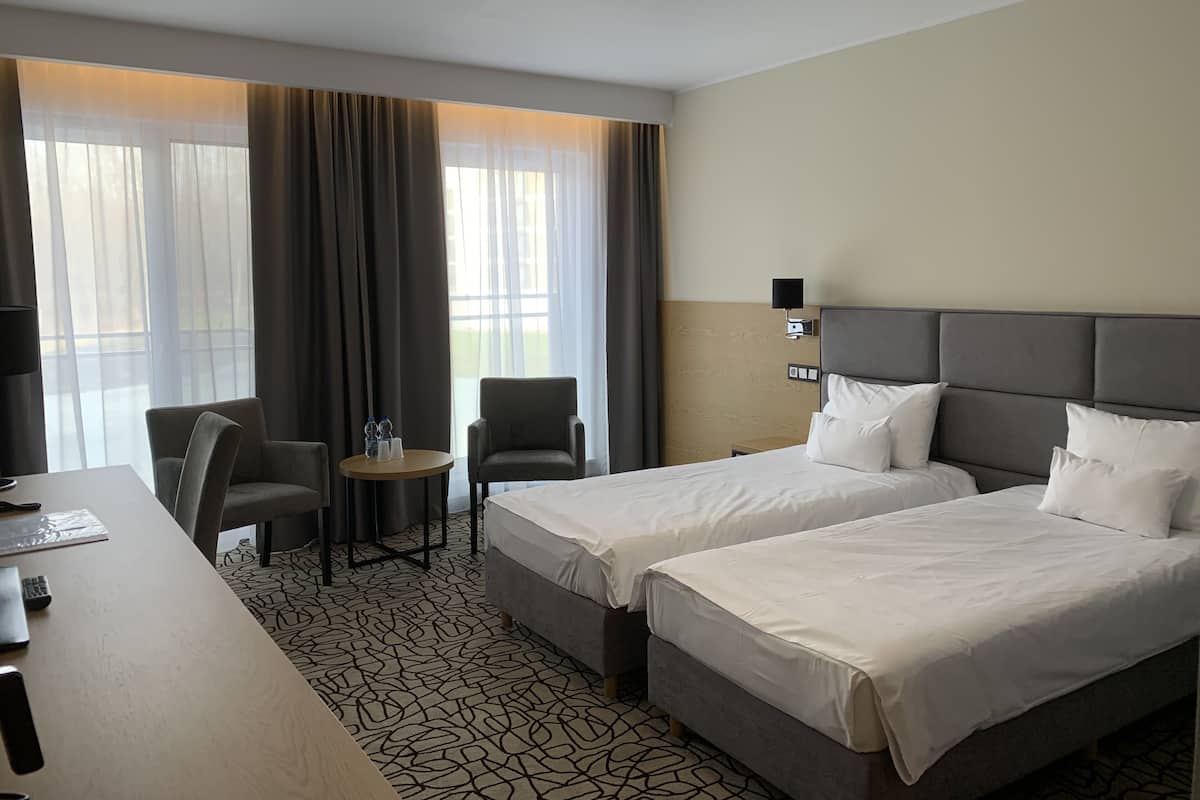 A hotel room with two beds, a desk, a chair, a small table with water bottles, and a lamp.