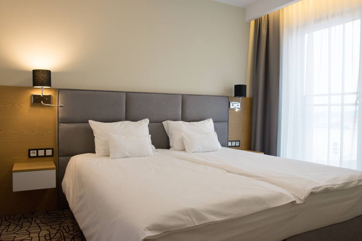 A hotel room with a large bed, two bedside tables, a wall-mounted lamp, and a window with curtains.