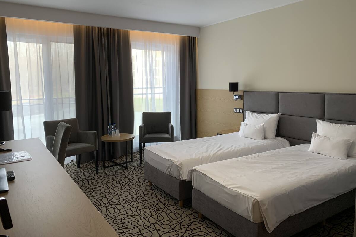 A hotel room with two beds, a desk, a chair, and a small table with water bottles.