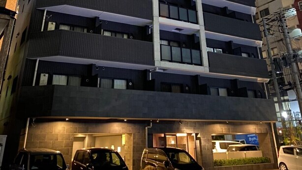 A multi-story building with a central staircase, multiple balconies, and a ground-level entrance with cars parked outside. - Local Hakata Apartment (Fukuoka)