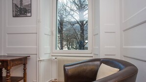 A room with a large window, a leather armchair, a wooden table, and a radiator. - Midi Guestrooms (Martigny)